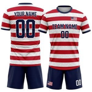 May include: A red and white striped soccer uniform with navy blue shorts. The jersey features the text "YOUR NAME" and "00" on the back, and "TEAM NAME" and "00" on the front. The American flag is also visible.