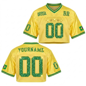 Puede incluir: Una camiseta amarilla recortada con detalles verdes, que muestra la palabra "BRASIL" y el n&uacute;mero "00" en purpurina verde. La camiseta tiene cuello en V y mangas cortas, con una bandera brasile&ntilde;a en la manga. La parte trasera de la camiseta dice "YOURNAME" y tiene el n&uacute;mero "00".