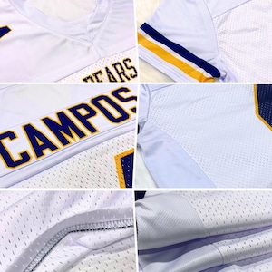 Puede incluir: Camiseta de f&uacute;tbol blanca con detalles azules y amarillos. La camiseta muestra las palabras "BEARS" y "CAMPOS" en letras azules. Tiene cuello en V y est&aacute; hecha de tejido de malla.