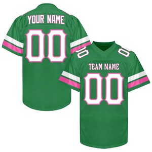 Puede incluir: Dos camisetas de f&uacute;tbol verdes con detalles blancos y rosas. Una muestra "YOUR NAME" y el n&uacute;mero "00" en blanco con contorno rosa. La otra muestra "TEAM NAME" y el n&uacute;mero "00".