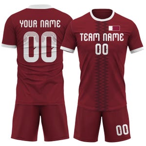 Op de afbeelding: Bordeaux voetbaltenue met witte accenten. Het shirt toont de tekst "TEAM NAME" en "00" in het wit, samen met een vlag van Qatar. De achterkant van het shirt toont "YOUR NAME" en "00". De shorts zijn bordeaux.
