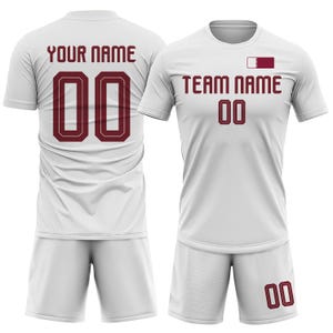 Op de afbeelding: Witte voetbaltenue met bordeauxrode accenten. Het shirt heeft "YOUR NAME" en het nummer "00" op de rug, en "TEAM NAME" en het nummer "00" op de voorkant. Inclusief bijpassende shorts.