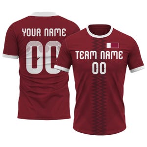 Op de afbeelding: Bordeauxrood voetbalshirt met witte randen en de tekst "TEAM NAME" en "00" op de voorkant. De achterkant van het shirt heeft de tekst "YOUR NAME" en "00". Het shirt heeft een vlag van Qatar op de voorkant.
