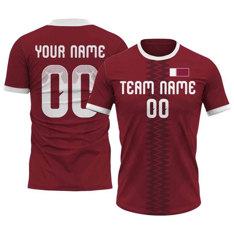 Op de afbeelding: Bordeauxrood voetbalshirt met witte randen en de tekst "TEAM NAME" en "00" op de voorkant. De achterkant van het shirt heeft de tekst "YOUR NAME" en "00". Het shirt heeft een vlag van Qatar op de voorkant.