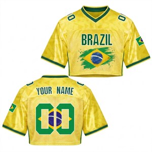 Puede incluir: Una camiseta corta amarilla con ribetes verdes y la palabra "BRAZIL" en letras verdes. La parte delantera presenta la bandera brasile&ntilde;a. La parte trasera tiene el texto "YOUR NAME" encima del n&uacute;mero "00" en verde. Una peque&ntilde;a bandera brasile&ntilde;a est&aacute; en la manga.