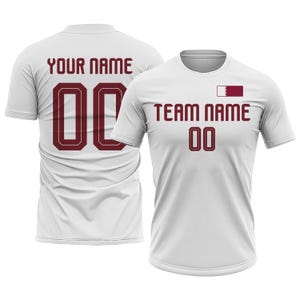 Op de afbeelding: Twee witte sportshirts met bordeauxrode tekst en cijfers. E&eacute;n shirt toont "YOUR NAME" en het nummer "00" op de achterkant. Het andere shirt heeft "TEAM NAME" en het nummer "00" op de voorkant, met een bordeauxrode en witte vlag.