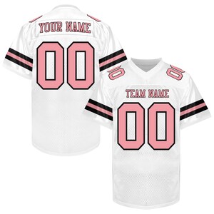 Puede incluir: Camiseta de f&uacute;tbol blanca con n&uacute;meros rosas y detalles negros. La camiseta tiene el n&uacute;mero "00" en la parte delantera y trasera, con "YOUR NAME" y "TEAM NAME" impresos en los hombros. Las mangas tienen rayas negras.