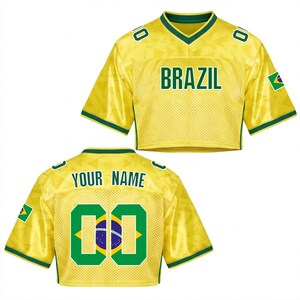 Puede incluir: Una camiseta amarilla recortada con ribetes verdes y la palabra "BRAZIL" en letras verdes. La camiseta tiene el n&uacute;mero "00" en la espalda con la bandera brasile&ntilde;a dentro de los n&uacute;meros. El texto "YOUR NAME" est&aacute; encima de los n&uacute;meros.