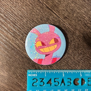 May include: A circular button featuring a cartoon character. The button has a pink character with a large yellow grin and the text "KAPTINKABLONEY". The button is approximately 5 cm in diameter.