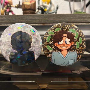 May include: Two round pinback buttons. One features a geometric design with holographic accents. The other has a cartoon illustration of a person with cucumbers and the word "cucumber" repeated around the edge. Both buttons are displayed on a clear plastic organizer.