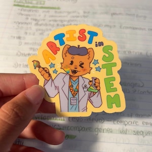 May include: A yellow sticker with the words "ARTIST in STEM" in colorful letters. The sticker features a cartoon fox character wearing a lab coat and holding a paintbrush and a beaker. The fox has a rainbow-colored cheek and is surrounded by blue stars.
