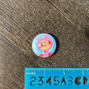 May include: A circular button with a cartoon character design. The button features a pink and yellow character with a wide grin, set against a blue background with pink accents. The button measures approximately 2.5 cm in diameter.