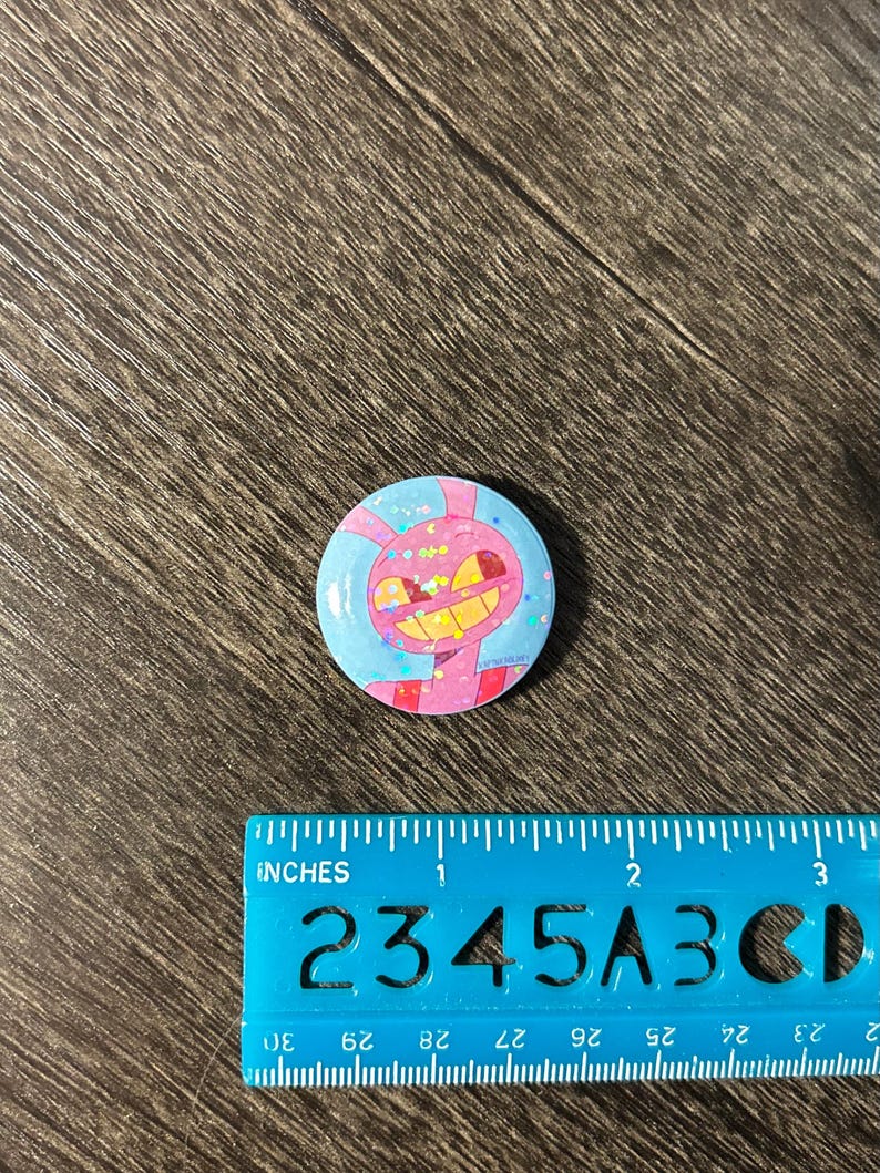 May include: A circular button with a cartoon character design. The button features a pink and yellow character with a wide grin, set against a blue background with pink accents. The button measures approximately 2.5 cm in diameter.