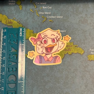 May include: A yellow sticker with a cartoon illustration of a duck with a pink bow, wearing a pink and blue shirt. The duck is smiling and has a yellow star above its head. The sticker is about 2 inches tall.