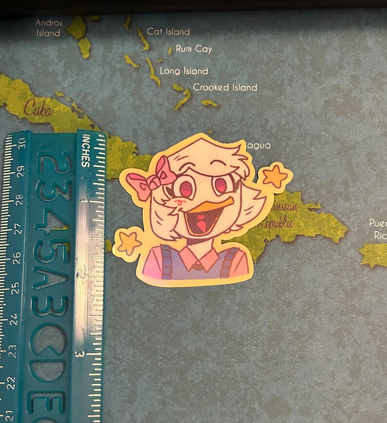 May include: A yellow sticker with a cartoon illustration of a duck with a pink bow, wearing a pink and blue shirt. The duck is smiling and has a yellow star above its head. The sticker is about 2 inches tall.