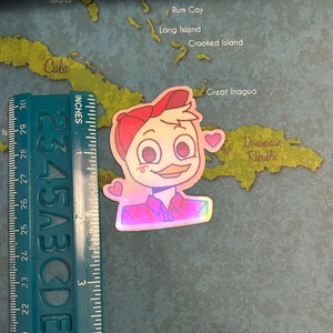 May include: A holographic sticker of a cartoon duck wearing a red baseball cap and a red and blue plaid shirt. The duck has a pink blush on its cheeks and is holding a pink heart in its hand. The sticker is on a map of the Caribbean.