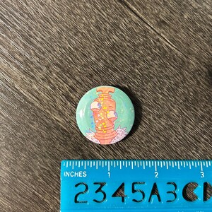 May include: A circular badge featuring a cartoon monster with three eyes and a pink body. The badge has a light blue background with glitter and is approximately 2.5 cm in diameter.