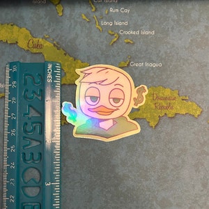 May include: A holographic sticker of a cartoon duck wearing a green shirt and holding a dollar bill. The duck has a light yellow beak and a light brown head. The sticker is on a map of the Caribbean.