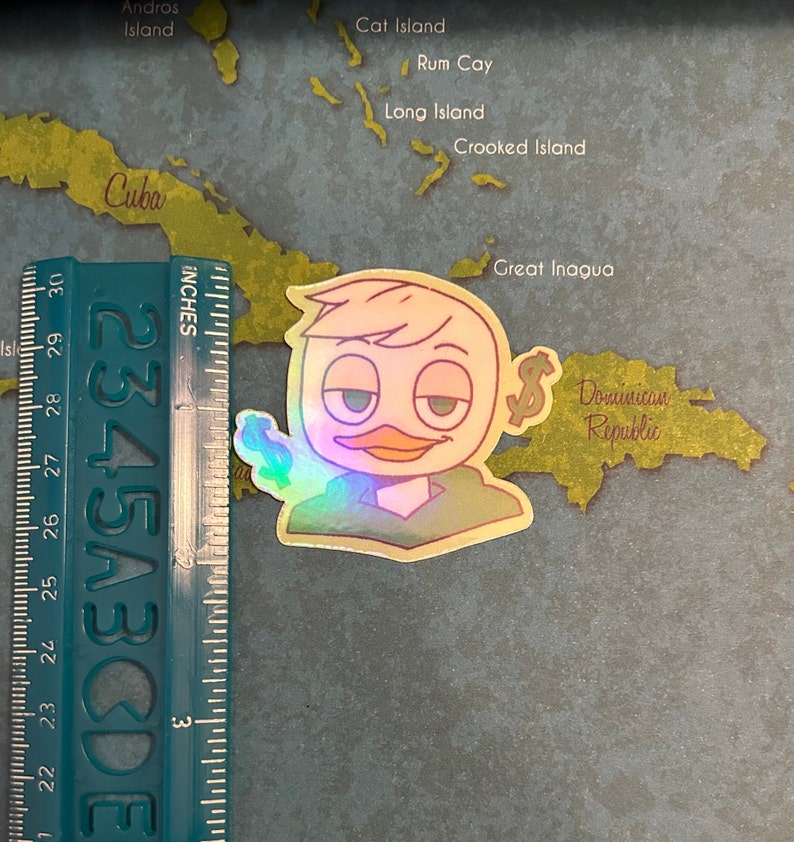 May include: A holographic sticker of a cartoon duck wearing a green shirt and holding a dollar bill. The duck has a light yellow beak and a light brown head. The sticker is on a map of the Caribbean.
