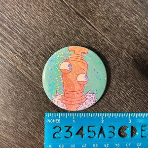 May include: A circular button featuring a cartoon character. The character is orange with large eyes and a purple collar, set against a teal background with glitter. The button is approximately 5 cm in diameter.