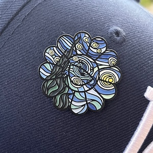 May include: A blue, green, and yellow enamel pin with a swirling, abstract design. The pin is attached to a dark blue baseball cap.