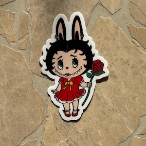May include: A die-cut sticker featuring a cartoon character with black hair and bunny ears, wearing a red dress and holding a red rose. The character has green eyes and a yellow bow. The sticker has a white border.