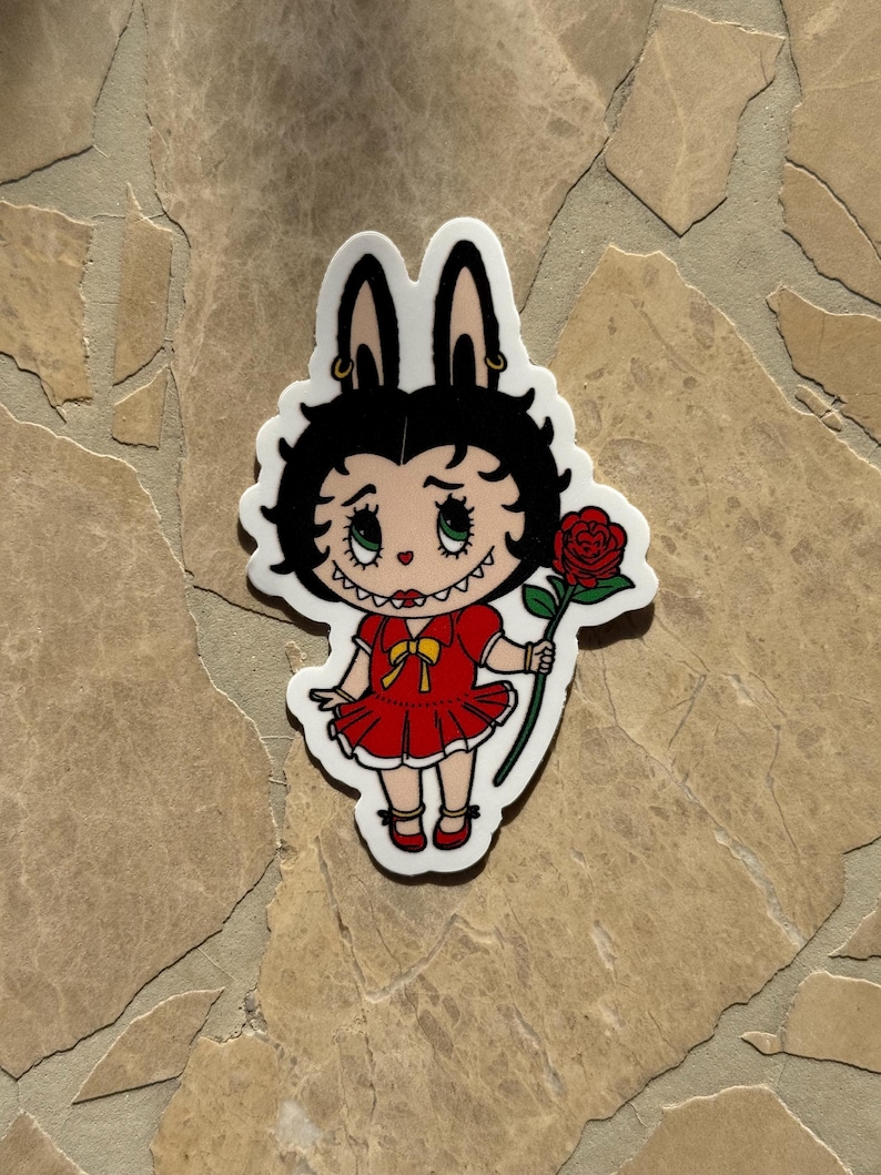 May include: A die-cut sticker featuring a cartoon character with black hair and bunny ears, wearing a red dress and holding a red rose. The character has green eyes and a yellow bow. The sticker has a white border.