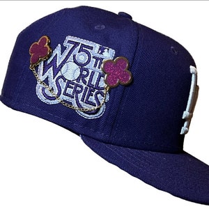May include: A navy blue baseball cap with a white '75th World Series' patch and two purple clover charms attached with a gold chain.