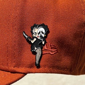 May include: A black and white enamel pin featuring a cartoon character holding a knife and wearing a Ghostface mask. The pin is on a brown baseball cap.