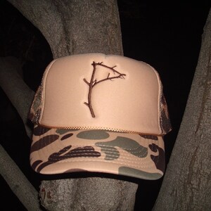 May include: A beige trucker hat with a camouflage brim. The hat has a brown embroidered design of a branch with a small flag on the end.