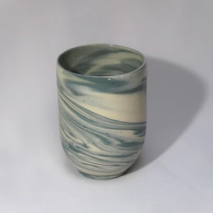 May include: A cylindrical ceramic cup featuring a marbled pattern of swirling blue-grey and off-white tones. The vessel has a smooth, rounded rim and a slightly tapered base, showcasing a fluid, blended aesthetic across the entire exterior surface.