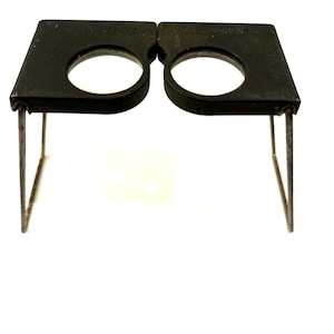 May include: A black, vintage jeweler's loupe with two circular lenses. The loupe has a rectangular frame and is supported by thin, metal legs. The lenses are clear, and the frame appears to be made of a dark, possibly painted, metal. The loupe is designed for close-up inspection.