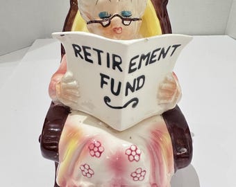 May include: A vintage ceramic cookie jar shaped like a seated elderly woman reading a newspaper. The woman has white hair, glasses, and is wearing a pink dress with floral accents. The newspaper reads "RETIREMENT FUND". The chair is brown and yellow.