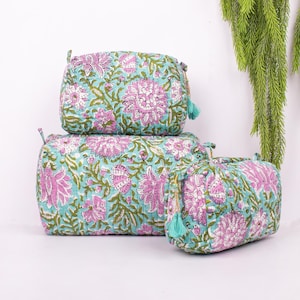 Peut inclure: Trois trousses de maquillage matelassées de tailles différentes, empilées. Elles présentent un motif floral avec des fleurs roses et des feuilles vertes sur un fond turquoise. Chaque trousse a un petit pompon et une fermeture éclair.