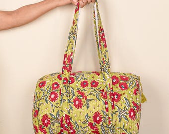 Puede incluir: Una bolsa de lona acolchada con estampado floral con flores rojas y hojas verdes sobre un fondo verde claro. La bolsa tiene dos asas largas y una borla amarilla. La bolsa está sujeta por una mano.