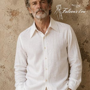 May include: A long-sleeved white button-down shirt with a pointed collar and a textured fabric finish. The shirt features a logo graphic in the upper right corner that includes a line drawing of a person and the text Falcon's Era.