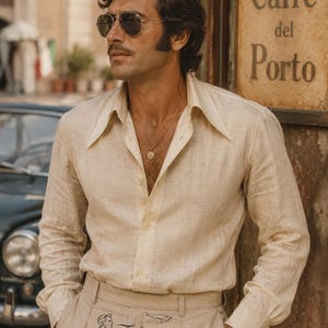 Puede incluir: Una camisa beige de manga larga con botones y cuello ancho, combinada con pantalones beige a juego que presentan un pequeño gráfico bordado de una persona y un pájaro sobre el texto Falcon's Era, usada con un collar con colgante dorado y gafas de sol.