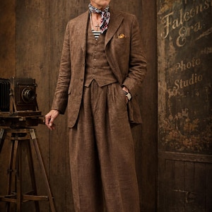 Può includere: Un completo in lino marrone, composto da blazer, gilet e pantaloni a gamba larga, indossato con basco e sciarpa. L'outfit è abbinato a scarpe marroni. Una macchina fotografica d'epoca su un treppiede e un'insegna con la scritta "Falcon's Era Photo Studio" sono sullo sfondo.