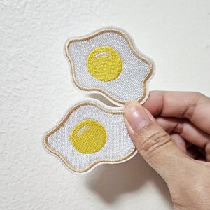 May include: Two white embroidered iron-on patches shaped like fried eggs with yellow yolks.