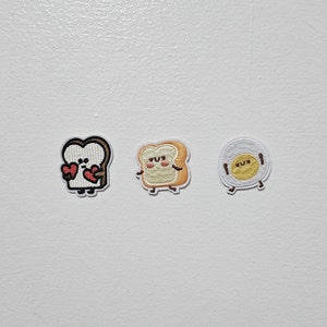 May include: Three embroidered patches featuring cartoon food characters. The first patch is a white bread slice with brown edges holding two red hearts. The second patch is a yellow and white bread slice with a brown crust. The third patch is a white egg with a yellow yolk.