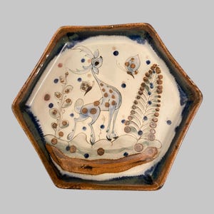 May include: Hexagonal ceramic plate with a hand-painted design. The plate features a deer with brown spots, a butterfly, and floral elements in blue, brown, and white. The rim has a brown and blue border.