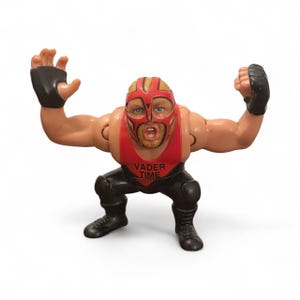 May include: A vintage wrestling action figure in a red and black outfit, with a red mask and the text "VADER TIME" on the chest. The figure is in a squatting pose with arms raised, wearing black gloves and boots.