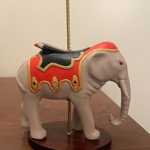 Może przedstawiać: Ceramiczna figurka słonia z karuzeli w kolorze jasnoszarym, z czerwono-czarnym siodłem i złotymi akcentami. Słoń ma trąbę i jest zamontowany na drewnianej podstawie ze złotym słupkiem i złotą kulą na górze.