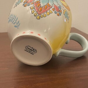 May include: A ceramic mug with a floral design in shades of blue, orange, and yellow. The mug has a light blue handle and a white base with pink polka dots. The bottom of the mug has the text "mudpie" and "MADE IN CHINA".