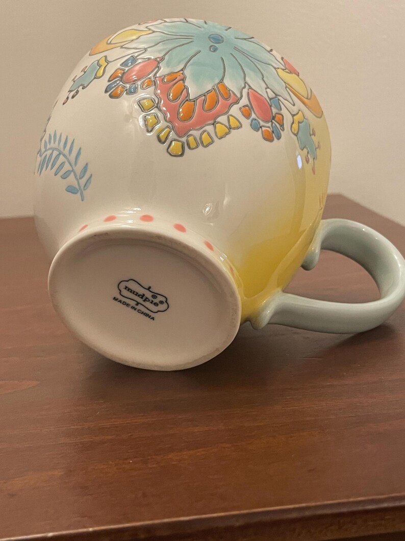 May include: A ceramic mug with a floral design in shades of blue, orange, and yellow. The mug has a light blue handle and a white base with pink polka dots. The bottom of the mug has the text "mudpie" and "MADE IN CHINA".