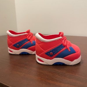 May include: A pair of miniature red sneakers with blue and white accents. The shoes have red laces, white soles, and blue details. The text "GP" is visible on the side of the shoe.