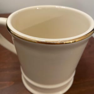 May include: A white ceramic mug with a gold rim. The mug has a rounded bottom and a handle.
