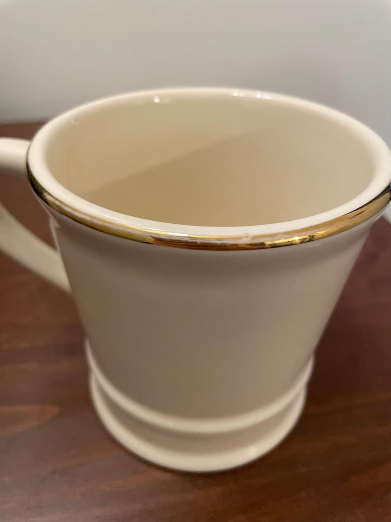 May include: A white ceramic mug with a gold rim. The mug has a rounded bottom and a handle.