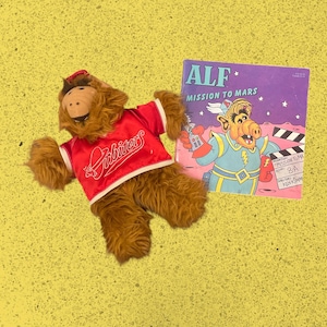 May include: A brown plush ALF doll wearing a red jersey with the word "Orbiters" in white script. Beside the doll is a comic book titled "ALF Mission to Mars" with a cartoon illustration of ALF.