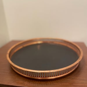 May include: A round copper-coloured serving tray with a black interior. The tray has a decorative, slotted rim. The tray is on a wooden surface.